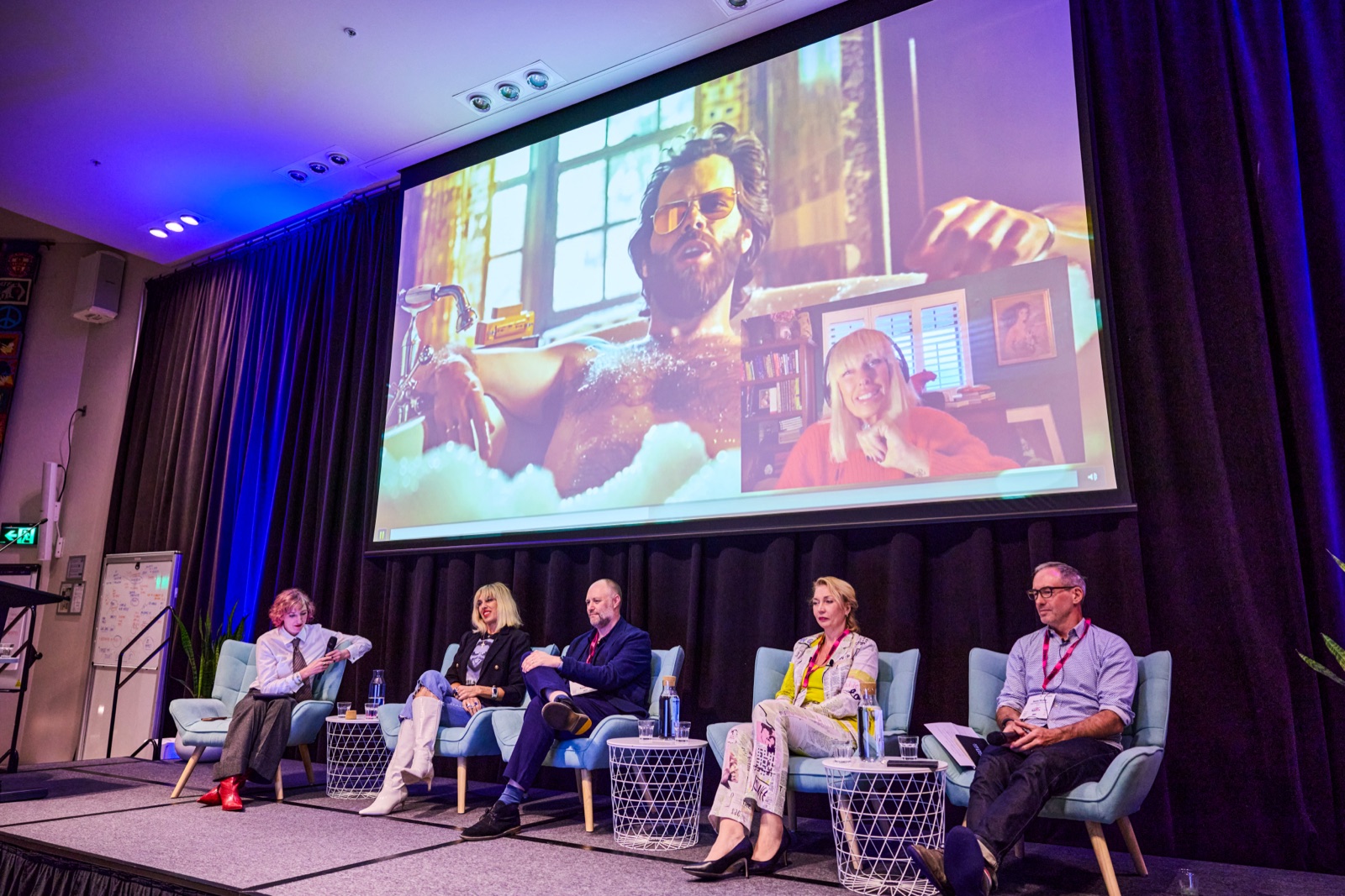 Five-person panel on stage with an AI-generated film clip playing on the large screen behind them