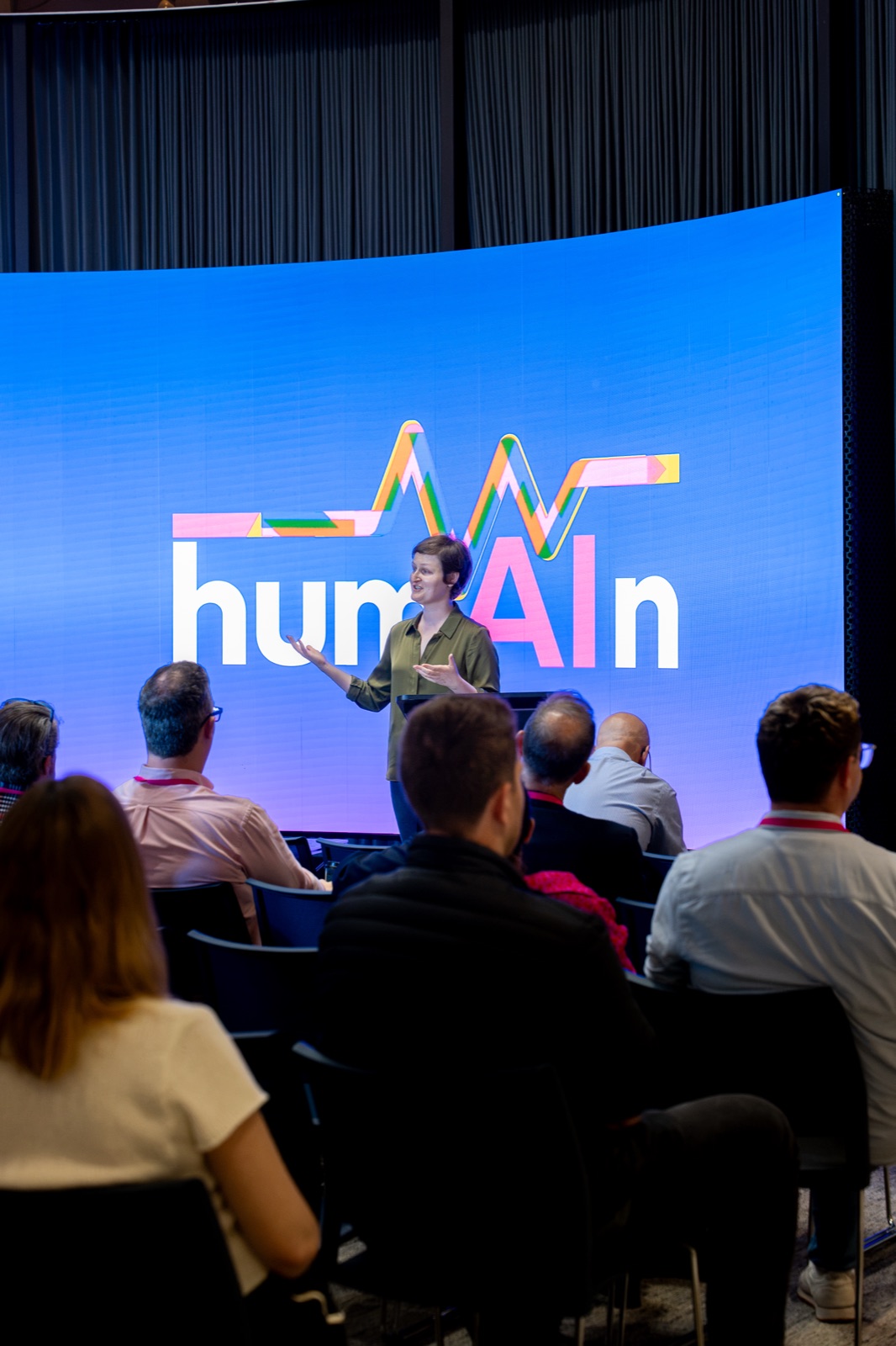 Speaker gesturing on stage in front of a large screen showing the humAIn logo, with the audience in the foreground