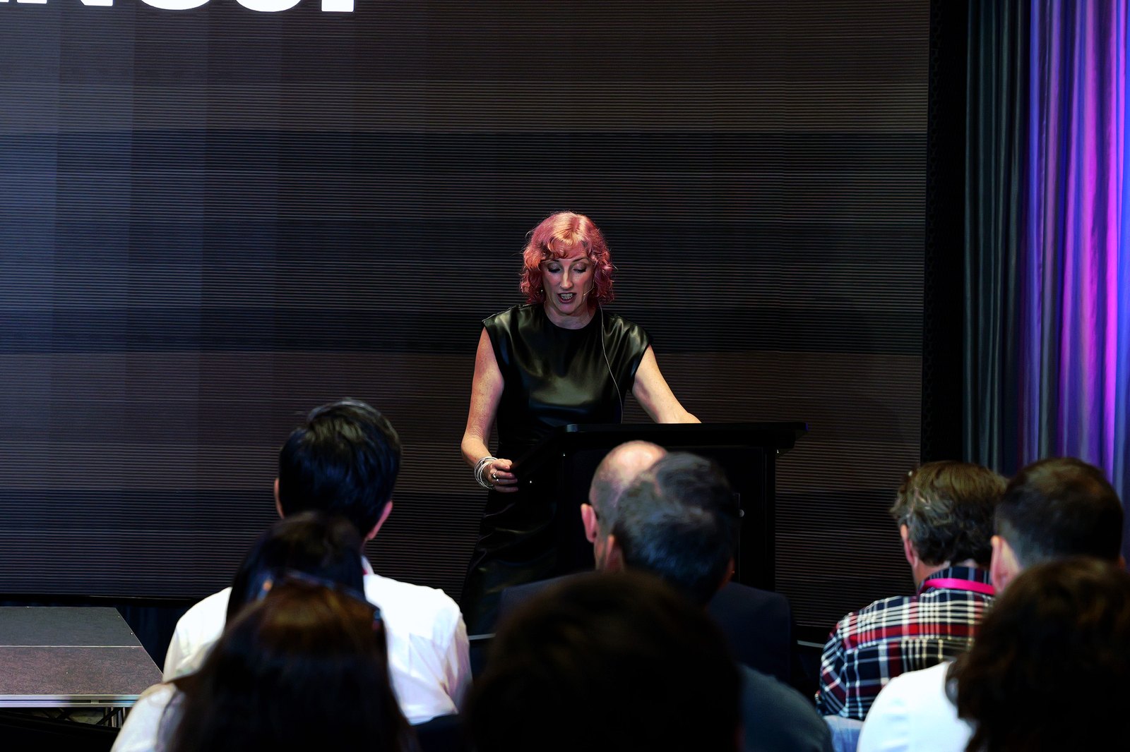 Cat McGinn at the lectern reading, in front of a dark humAIn screen with the audience in the foreground
