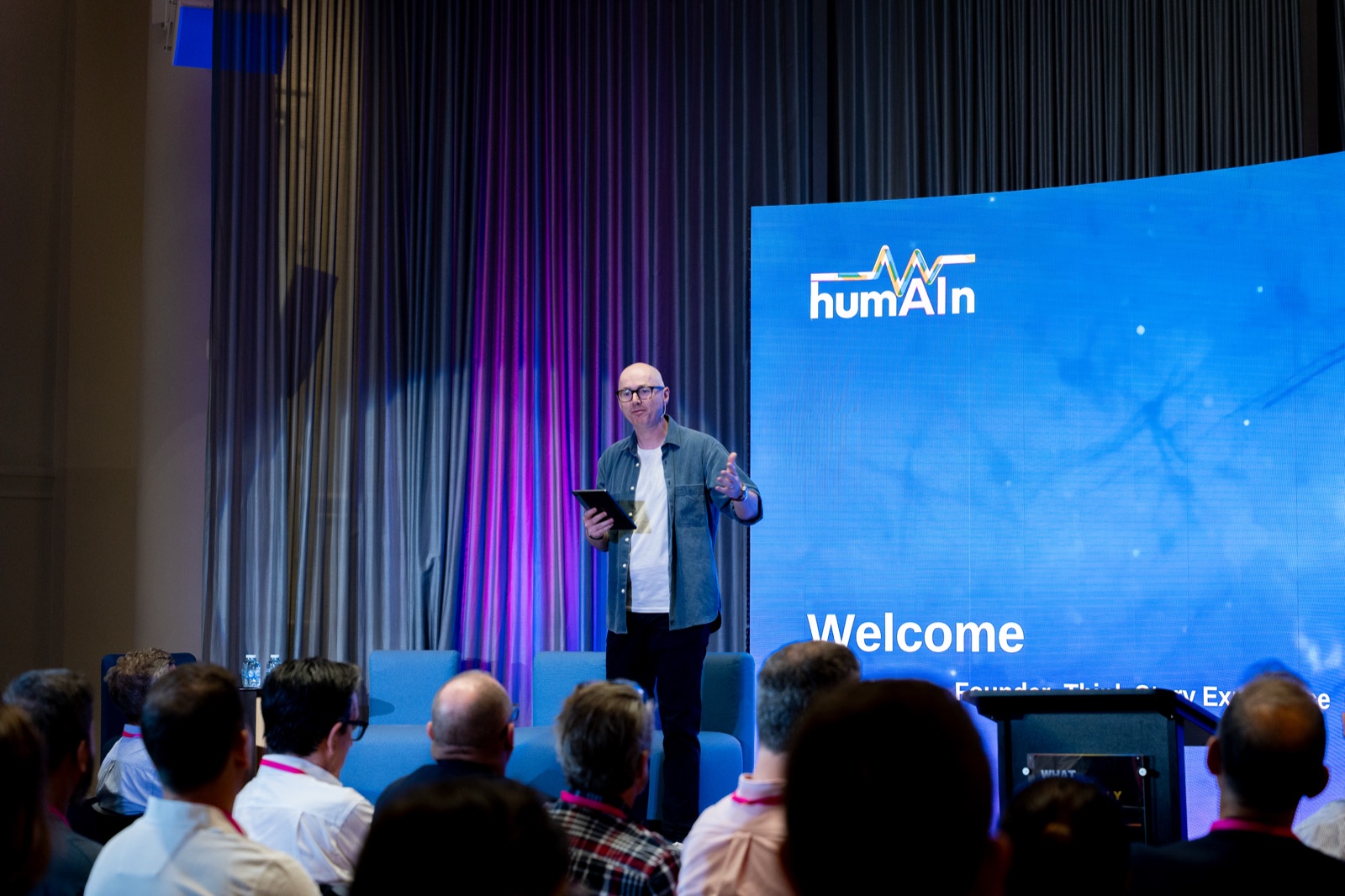 Speaker holding a tablet on stage beside a screen reading Welcome, with the audience in the foreground