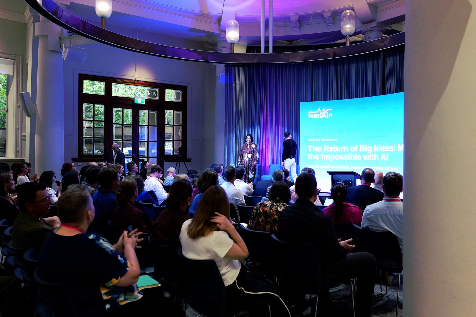 Wide view of the room with a speaker on stage beside a slide titled "The Return of Big Ideas"