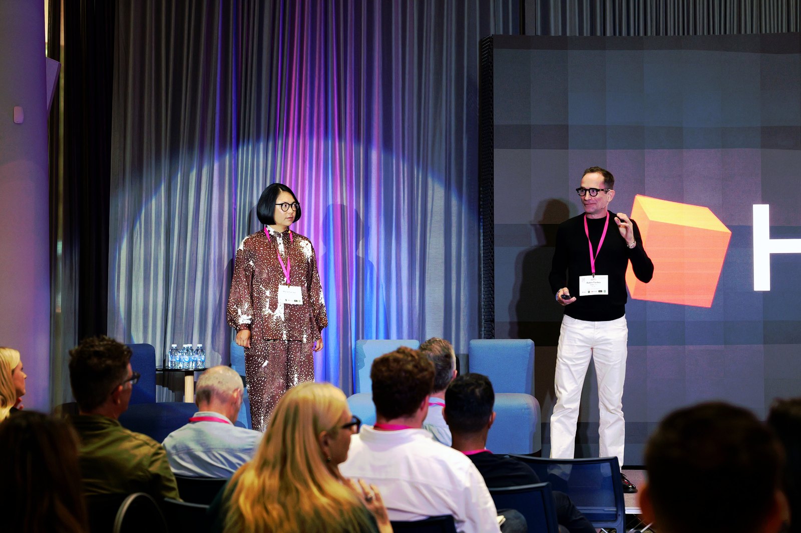 A woman in a patterned suit and a man in black with white trousers speaking on stage