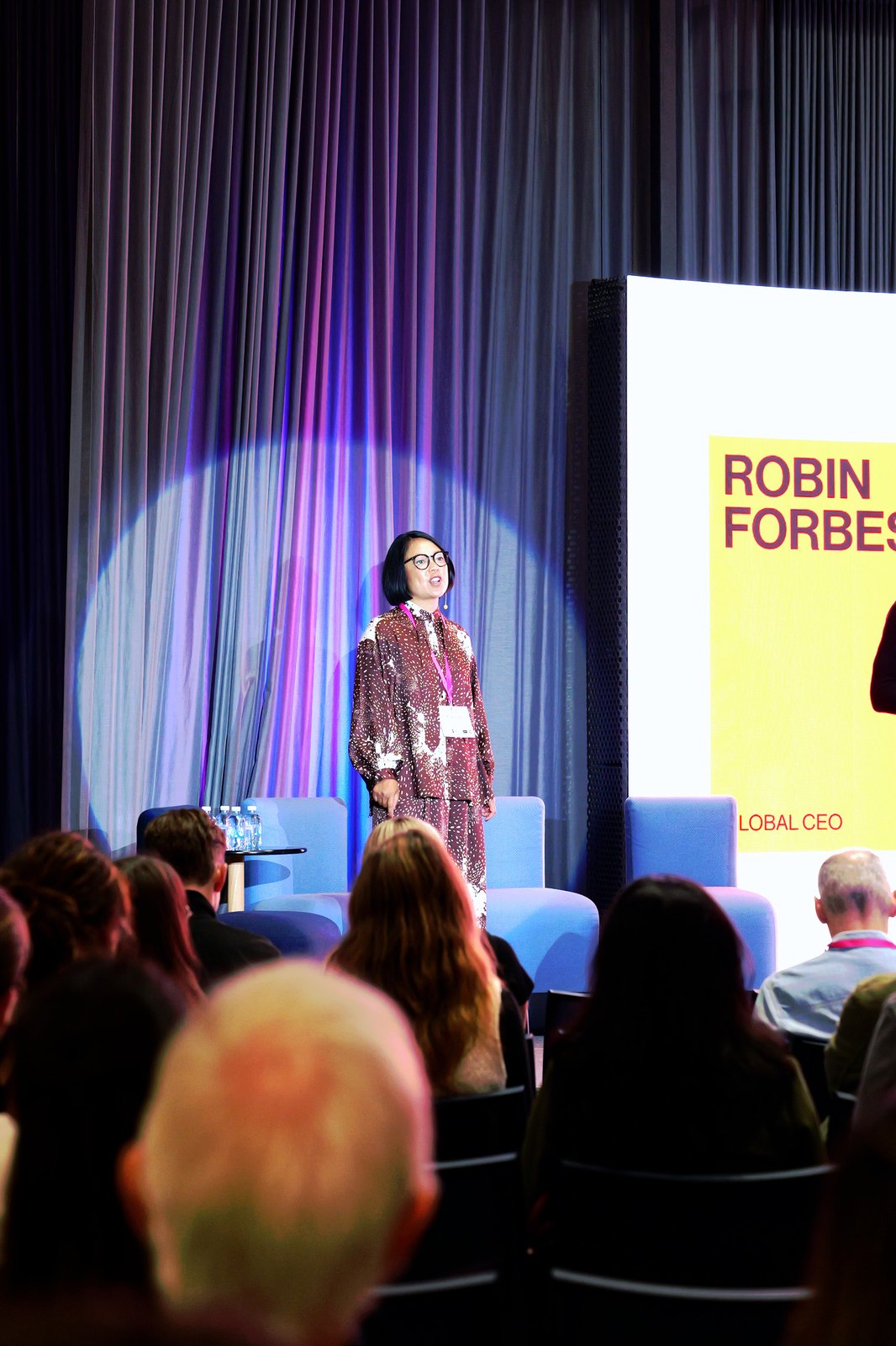 A woman in a patterned suit on stage beside a slide naming Robin Forbes