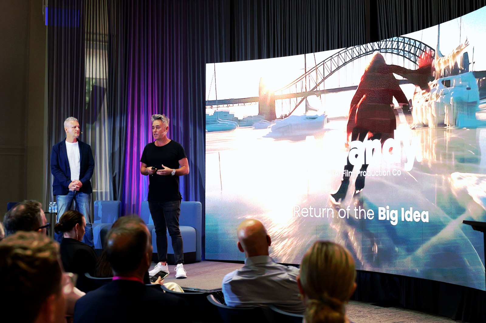Two men on stage in front of a screen showing "AiCandy - Return of the Big Idea"