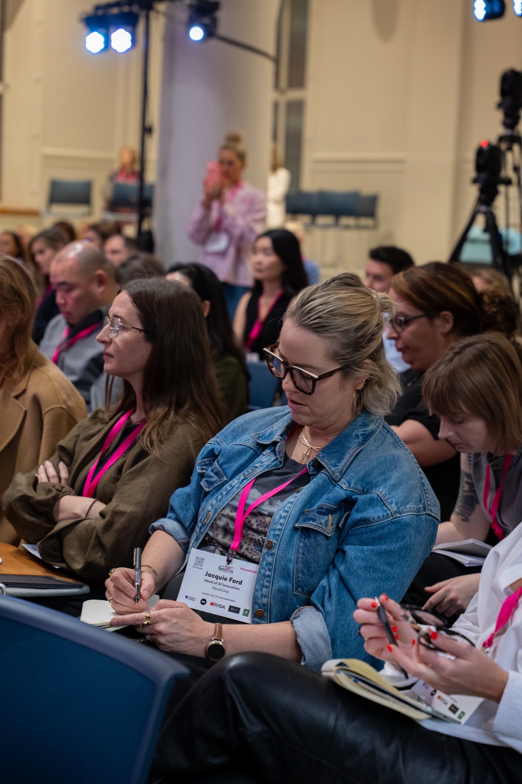 Delegates seated in the audience wearing pink lanyards, one taking notes