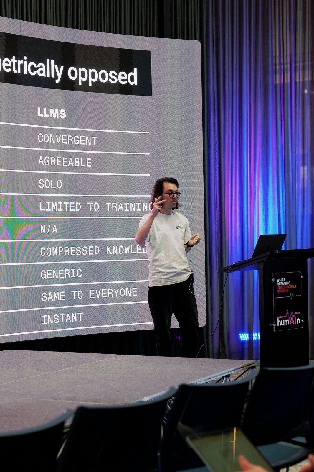A speaker in a white t-shirt at the lectern beside a slide comparing LLMs