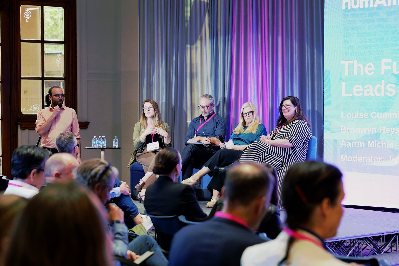 A panel on stage in front of a humAIn slide listing Louise Cummins, Bronwyn Heys and Aaron Michie as panellists and Joana Barros as moderator