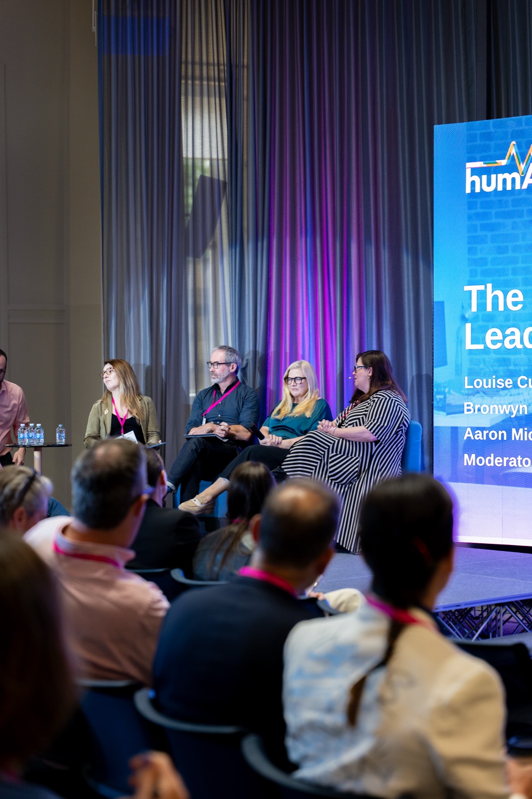 Panel of speakers seated on stage beside a large screen, with the audience in the foreground