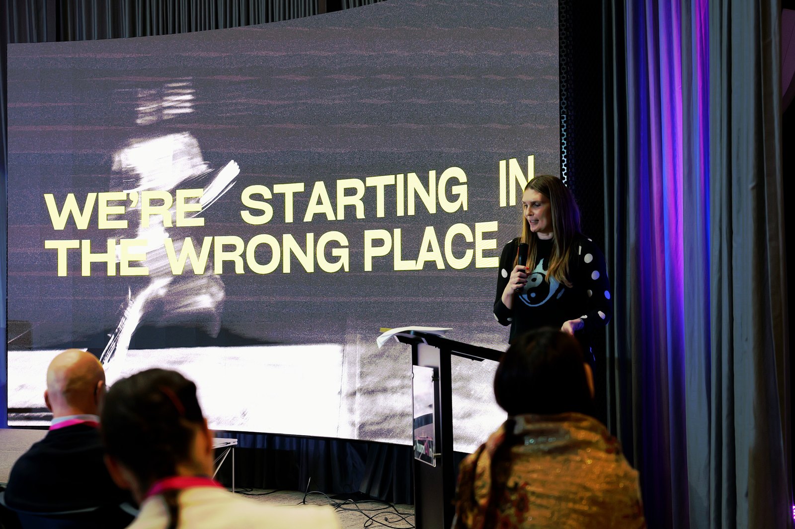A woman in a spotted dress speaking at the lectern in front of a slide reading "We're starting in the wrong place"