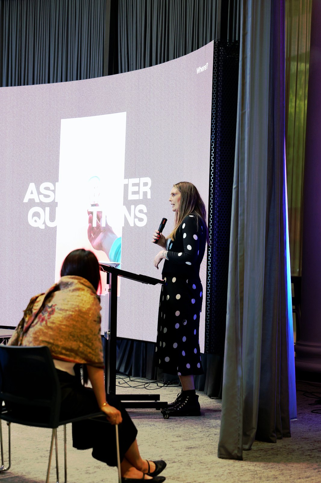 A woman in a spotted dress speaking in front of a slide about audience questions