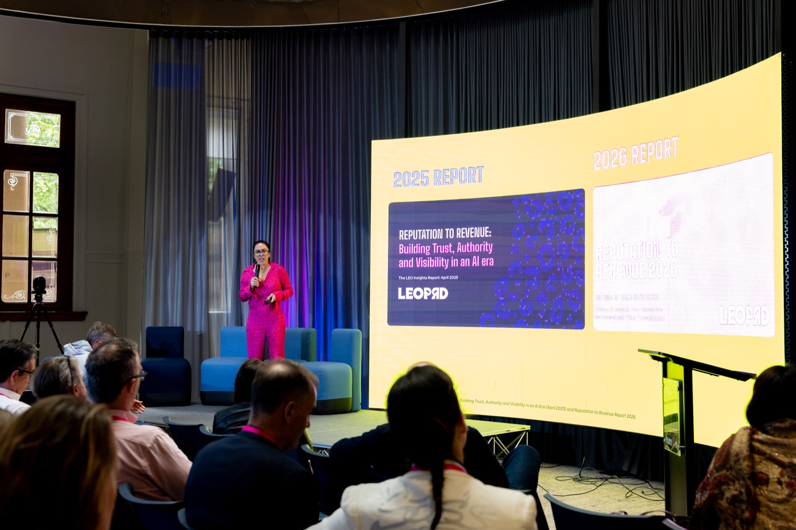 Speaker in a bright pink suit presenting on stage beside a screen showing a report cover
