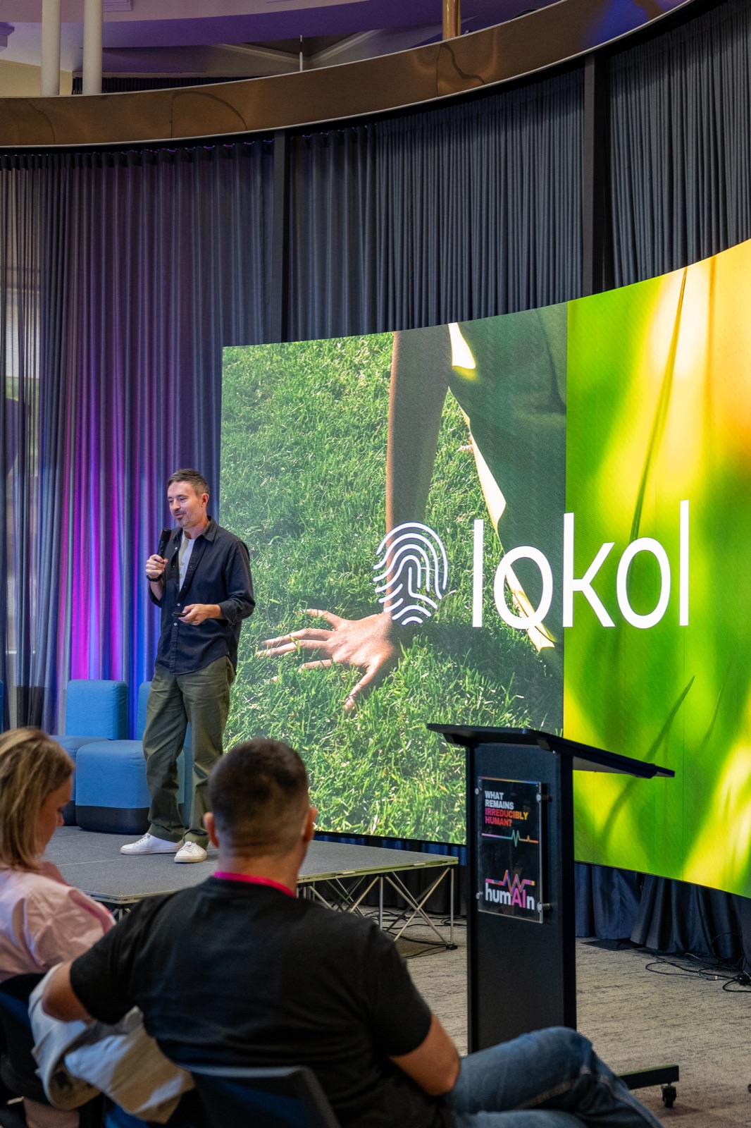 Speaker holding a microphone on stage in front of a screen showing a logo over a close-up of grass