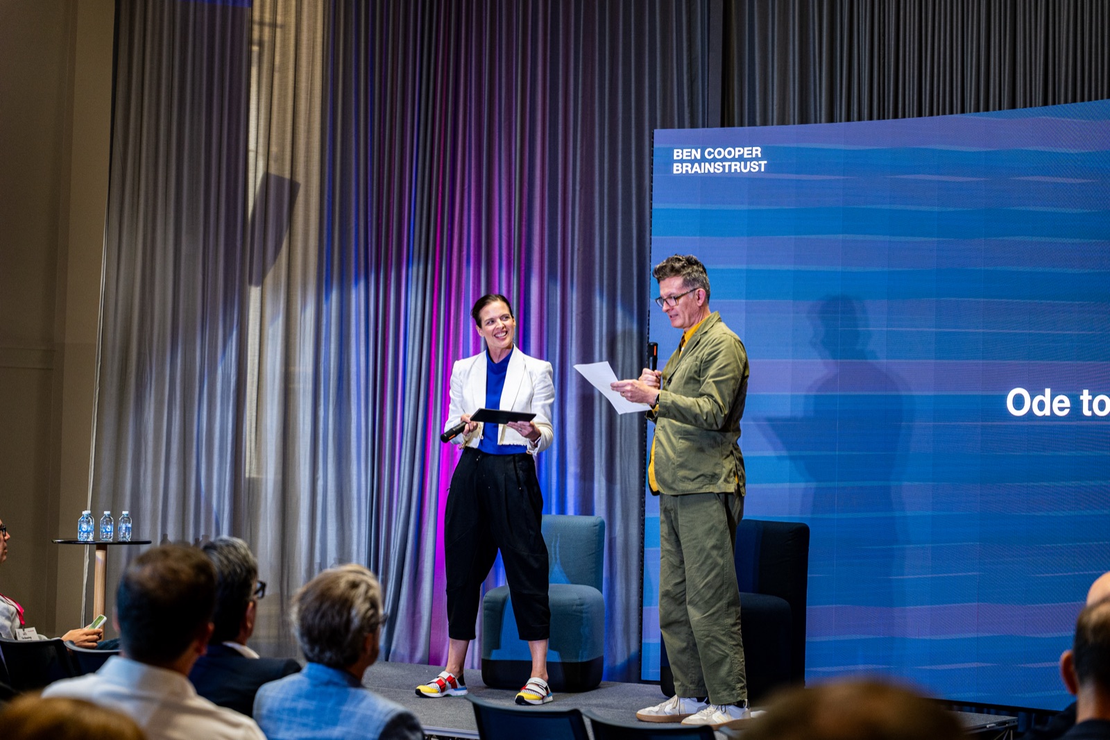 Two people on stage in front of a blue screen, one holding a sheet of paper
